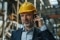 Person mit gelbem Schutzhelm spricht am Smartphone in einer industriellen Umgebung mit Maschinen im Hintergrund.
